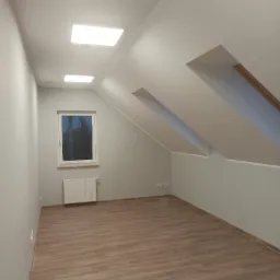 Poddasze po remoncie: świeżo pomalowane szare ściany, jasne panele podłogowe, biała rama okienna z widokiem na drzewa, grzejnik pod oknem, wbudowane kwadratowe lampy sufitowe.