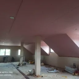 Montaż płyt na poddaszu, widoczne płyty gipsowo-kartonowe na skośnym suficie, pracownicy na drabinie i bałagan na podłodze.