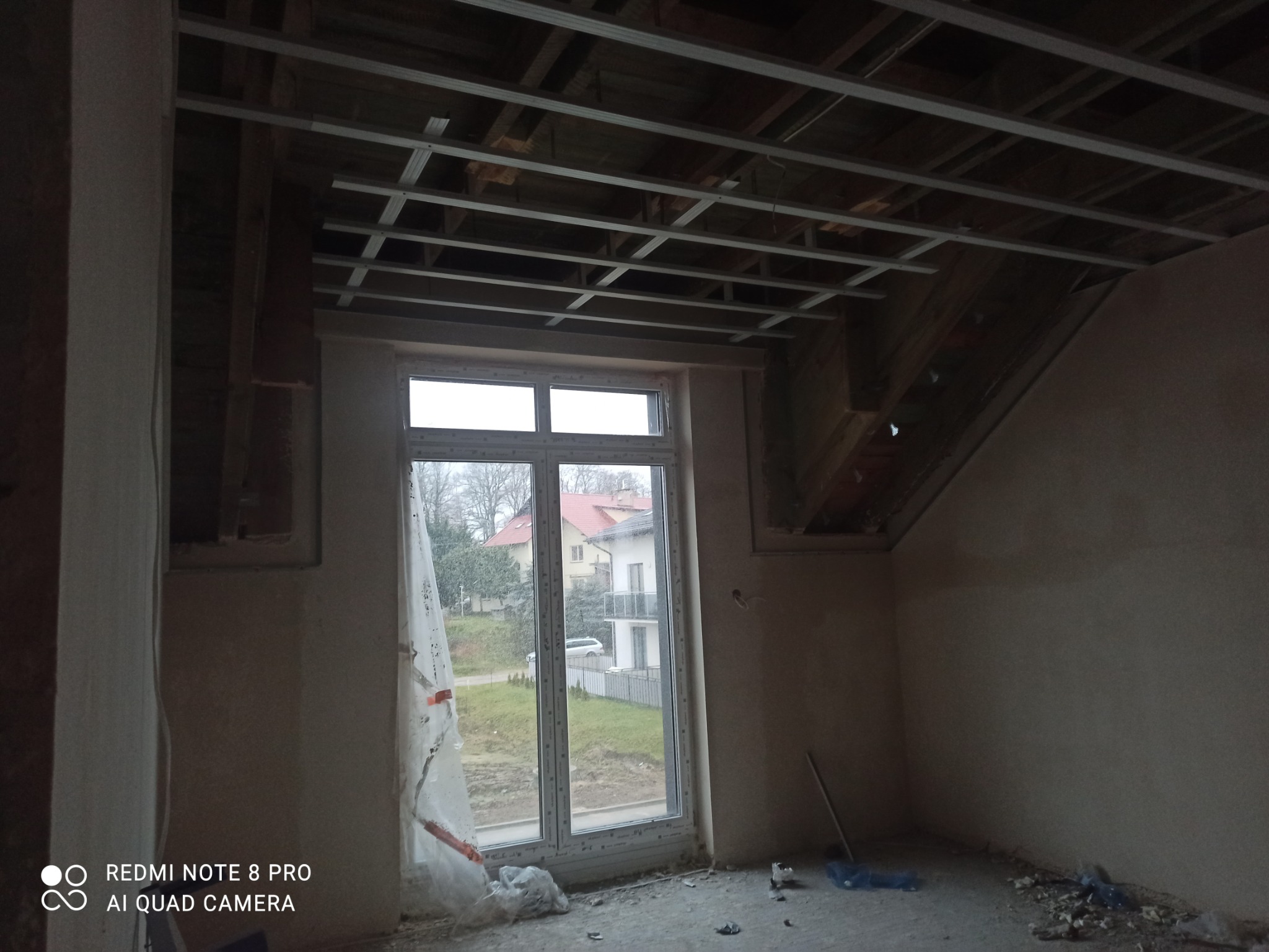 Metalowa konstrukcja podwieszanego sufitu zamontowana w pokoju na poddaszu, widoczne drewniane elementy dachu i okno z widokiem na domy.