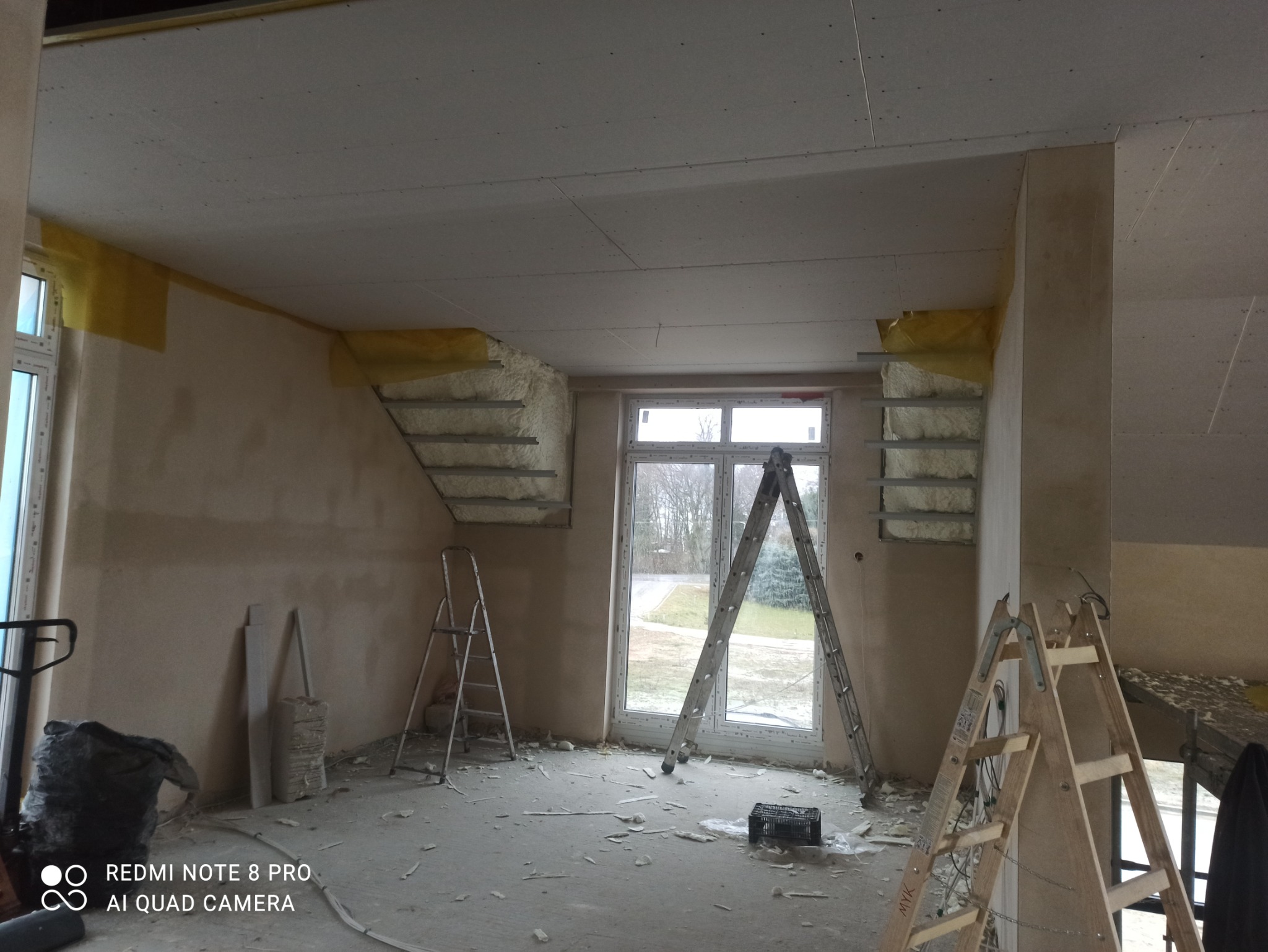 Montaż płyty karton gips na suficie poddasza, widoczne profile i ocieplenie wełną mineralną, dwie drabiny rozstawione w pomieszczeniu, okno z widokiem na drzewa.