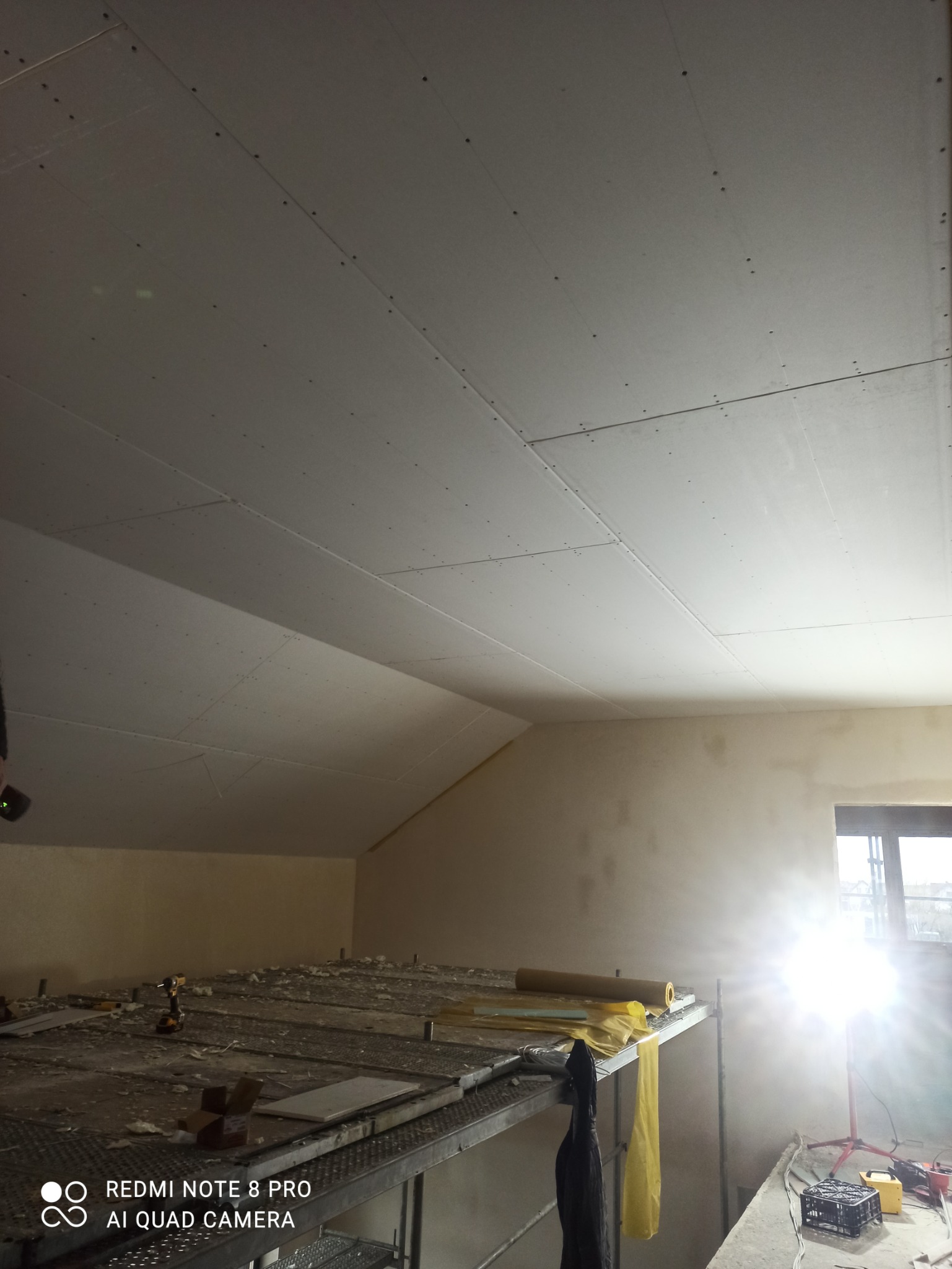 Montaż sufitu z płyt gipsowo-kartonowych na poddaszu, widoczne łączenia płyt i śruby, rusztowanie z narzędziami w tle.