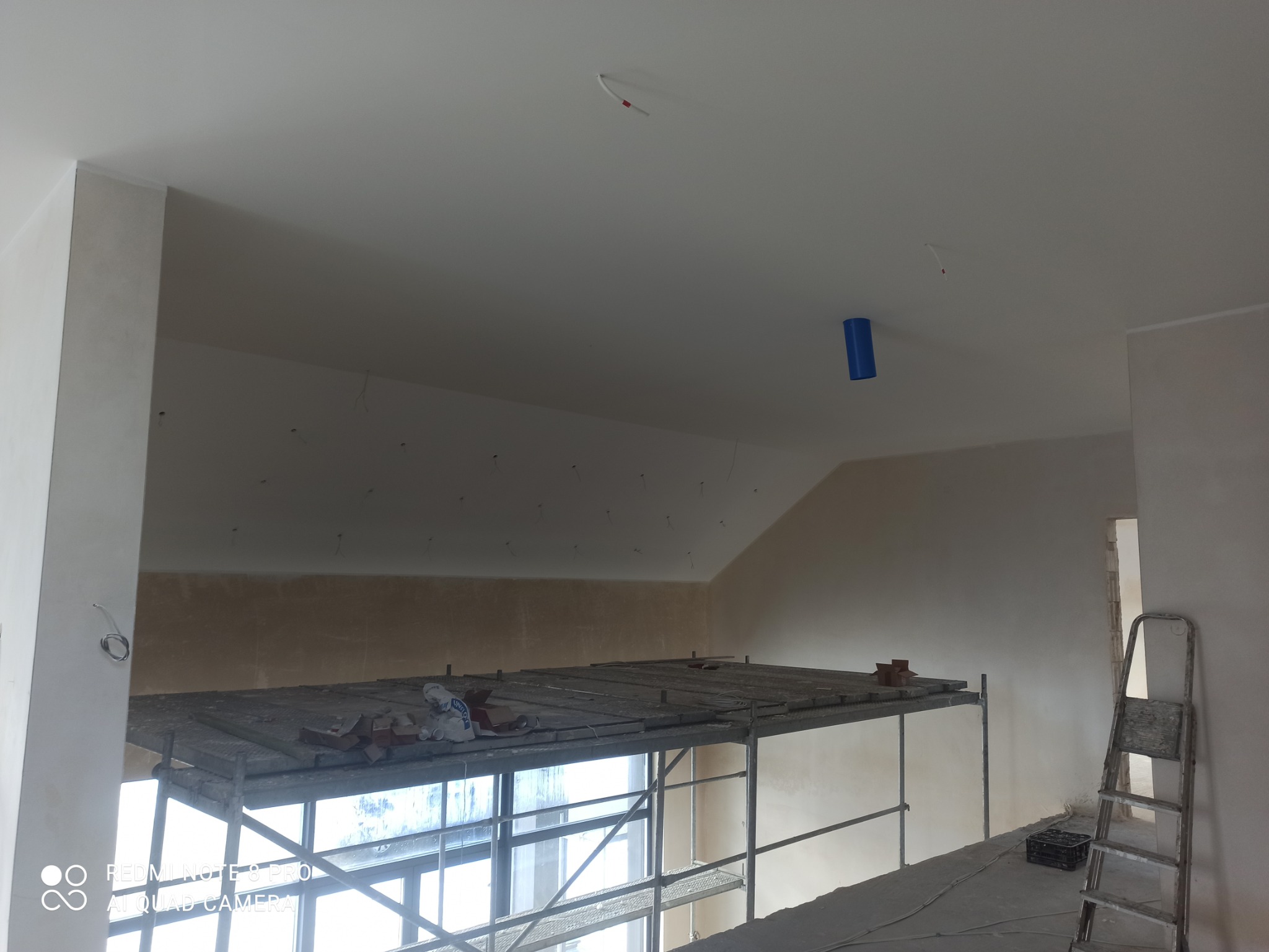 Wnętrze w trakcie remontu: rusztowanie z cegłami i workami, ściany przygotowane do malowania, sufit z wystającymi kablami i niebieską rurką, drabina oparta o ścianę.