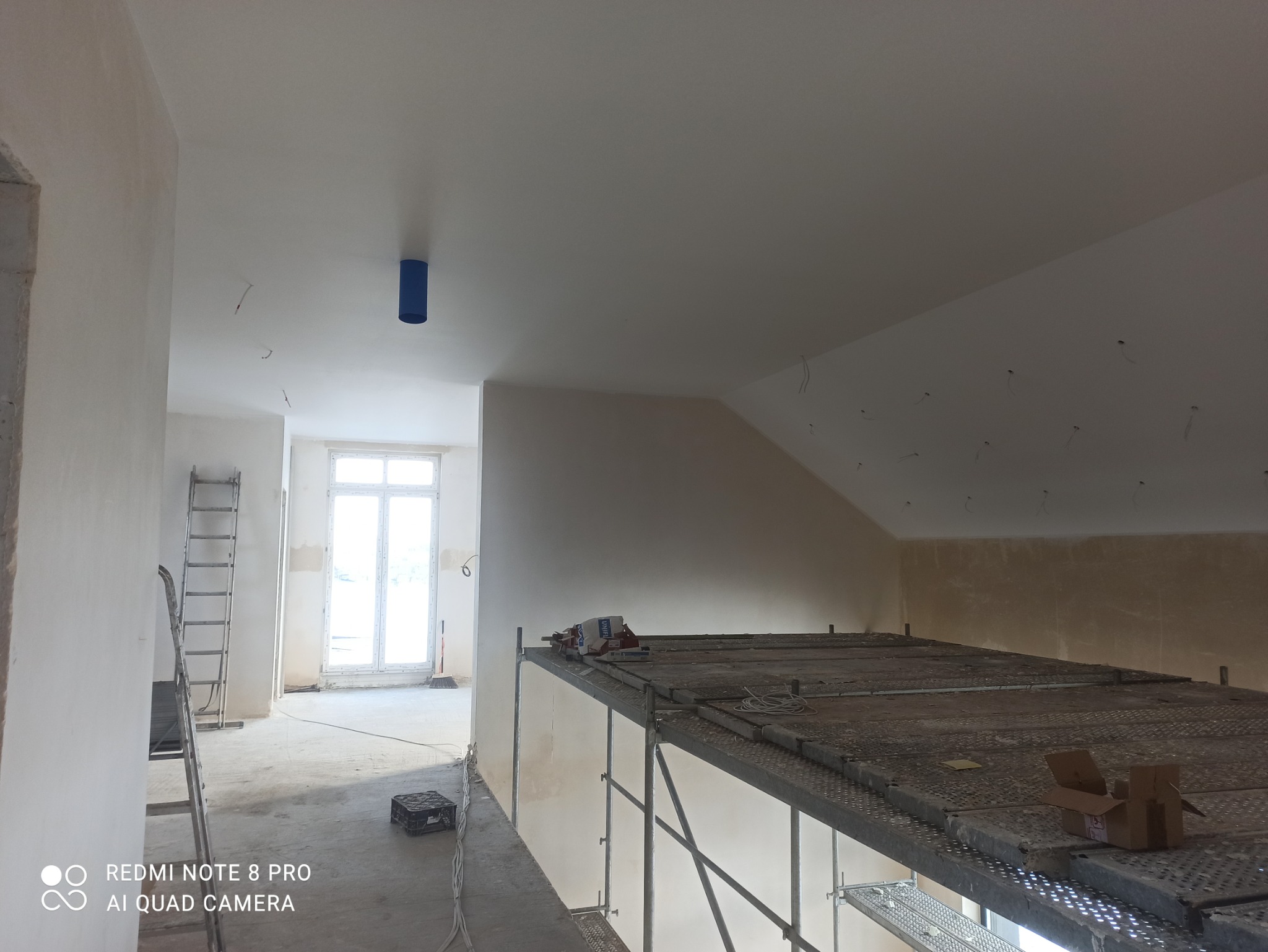 Wnętrze w trakcie remontu: widok z rusztowania na świeżo zagruntowane ściany i sufit z wystającymi przewodami elektrycznymi, w tle okno i drabina oparta o ścianę.