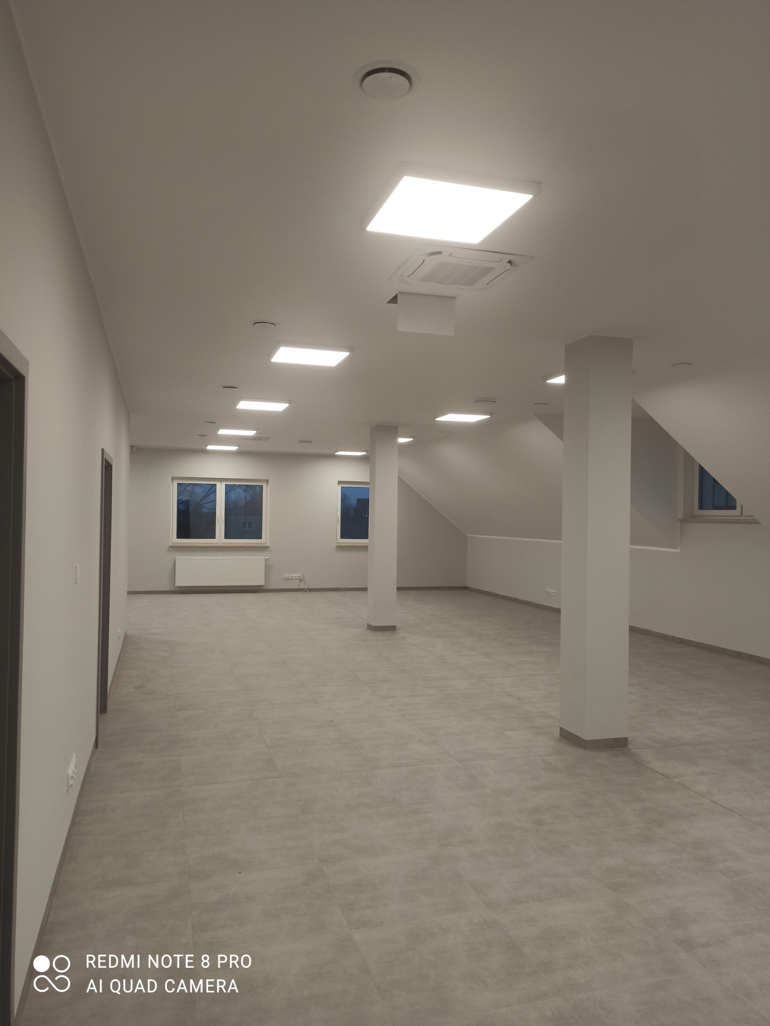 Wykończone wnętrze biurowe z szarą podłogą z płytek, białymi ścianami, kolumnami i oknami z grzejnikami, oświetlone kwadratowymi lampami sufitowymi i klimatyzacją.