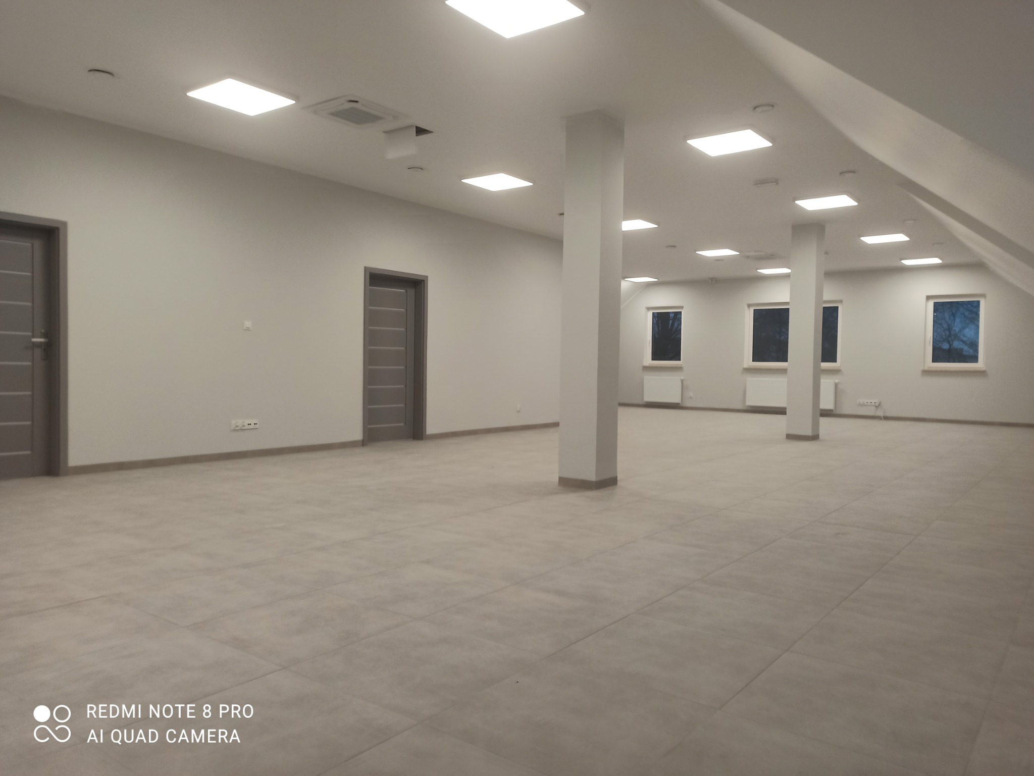 Przestronne, jasne wnętrze biurowe z szarymi drzwiami, kolumnami i kwadratowymi lampami sufitowymi, widok na zewnątrz przez okna.