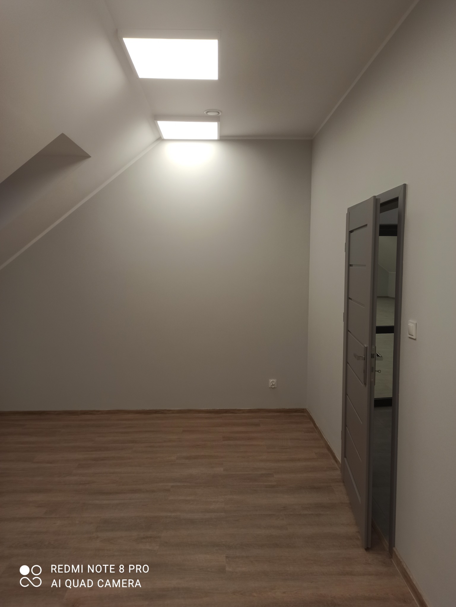 Wnętrze pokoju na poddaszu po remoncie: szare ściany, drewnopodobne panele podłogowe, otwarte szare drzwi, dwa kwadratowe panele LED w suficie.
