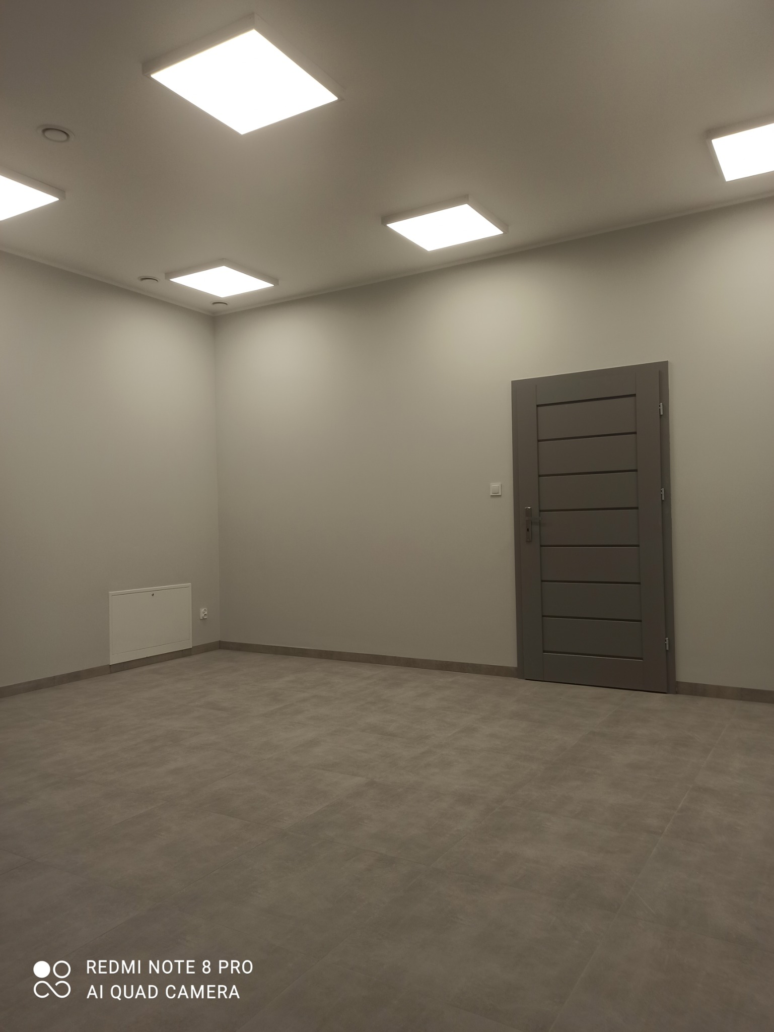 Puste pomieszczenie po remoncie, szare płytki podłogowe, szare ściany, szara, nowoczesna stolarka drzwiowa, białe, kwadratowe lampy sufitowe LED.