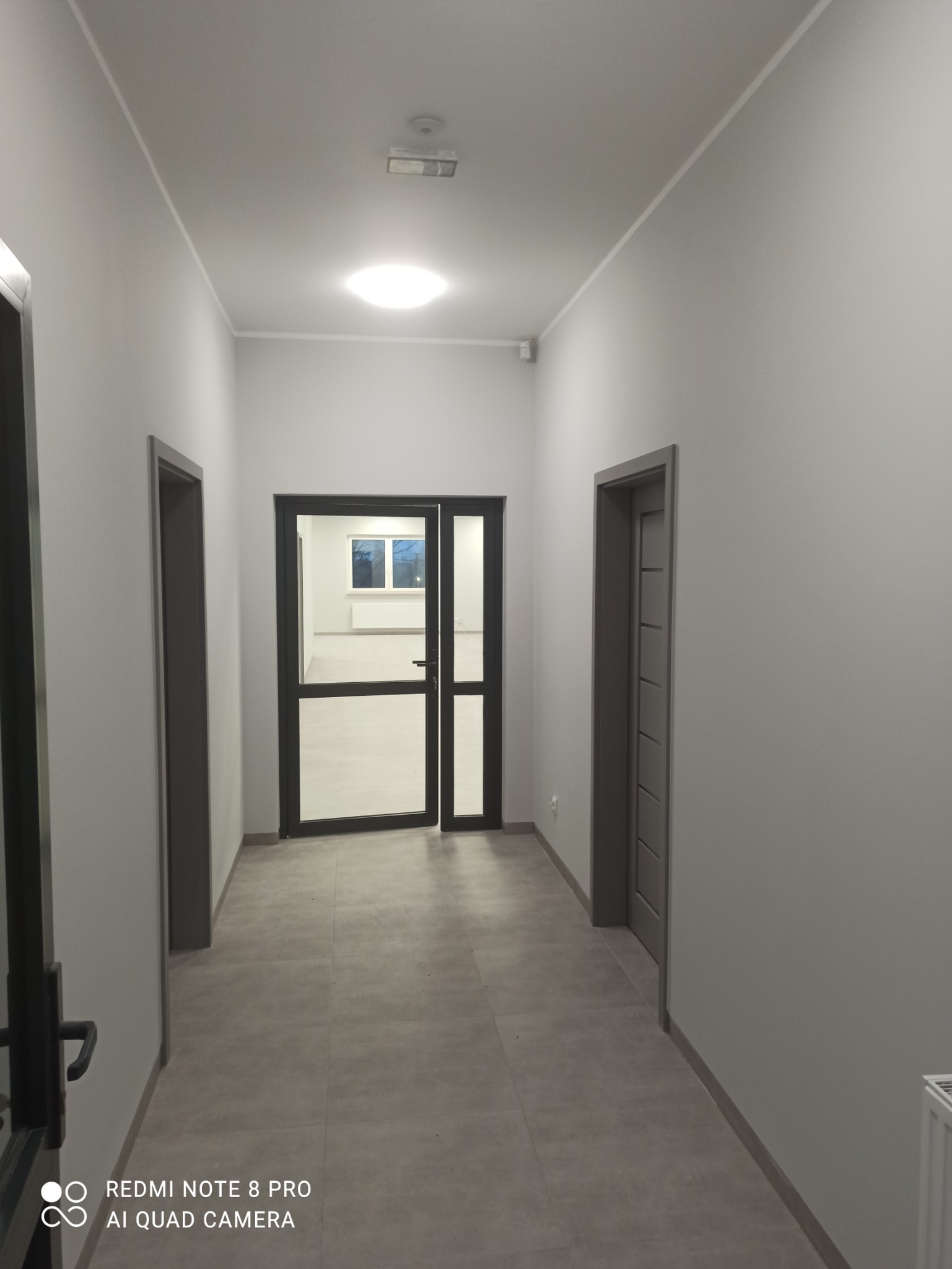 Długi korytarz w biurze lub budynku użyteczności publicznej, świeżo po remoncie, z szarymi ścianami, szarą podłogą z płytek i szarymi drzwiami, widok na przeszklone drzwi na końcu korytarza.