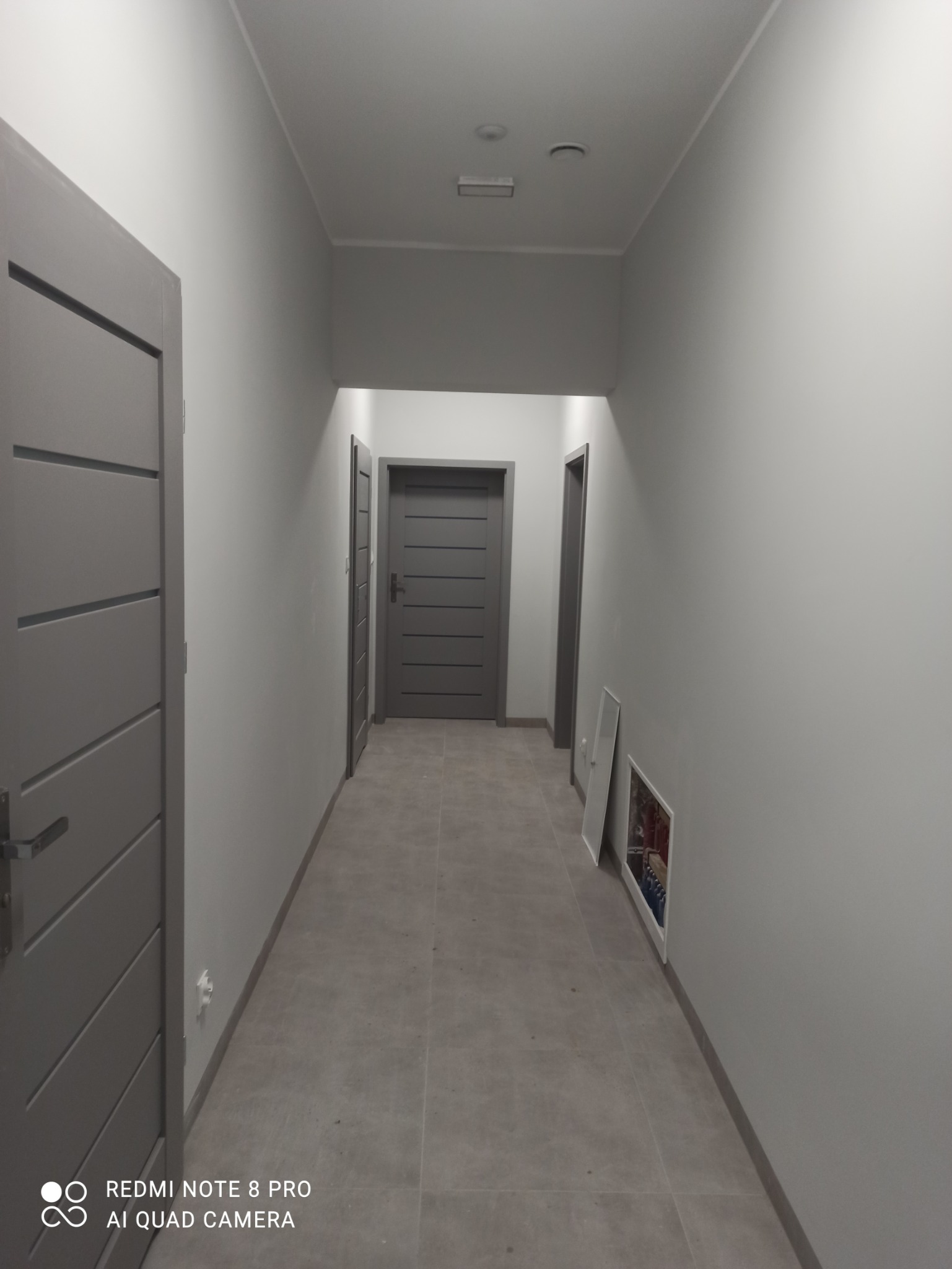 Szary korytarz z trzema parami drzwi, wykończony w minimalistycznym stylu, z płytkami na podłodze i obrazem opartym o ścianę.