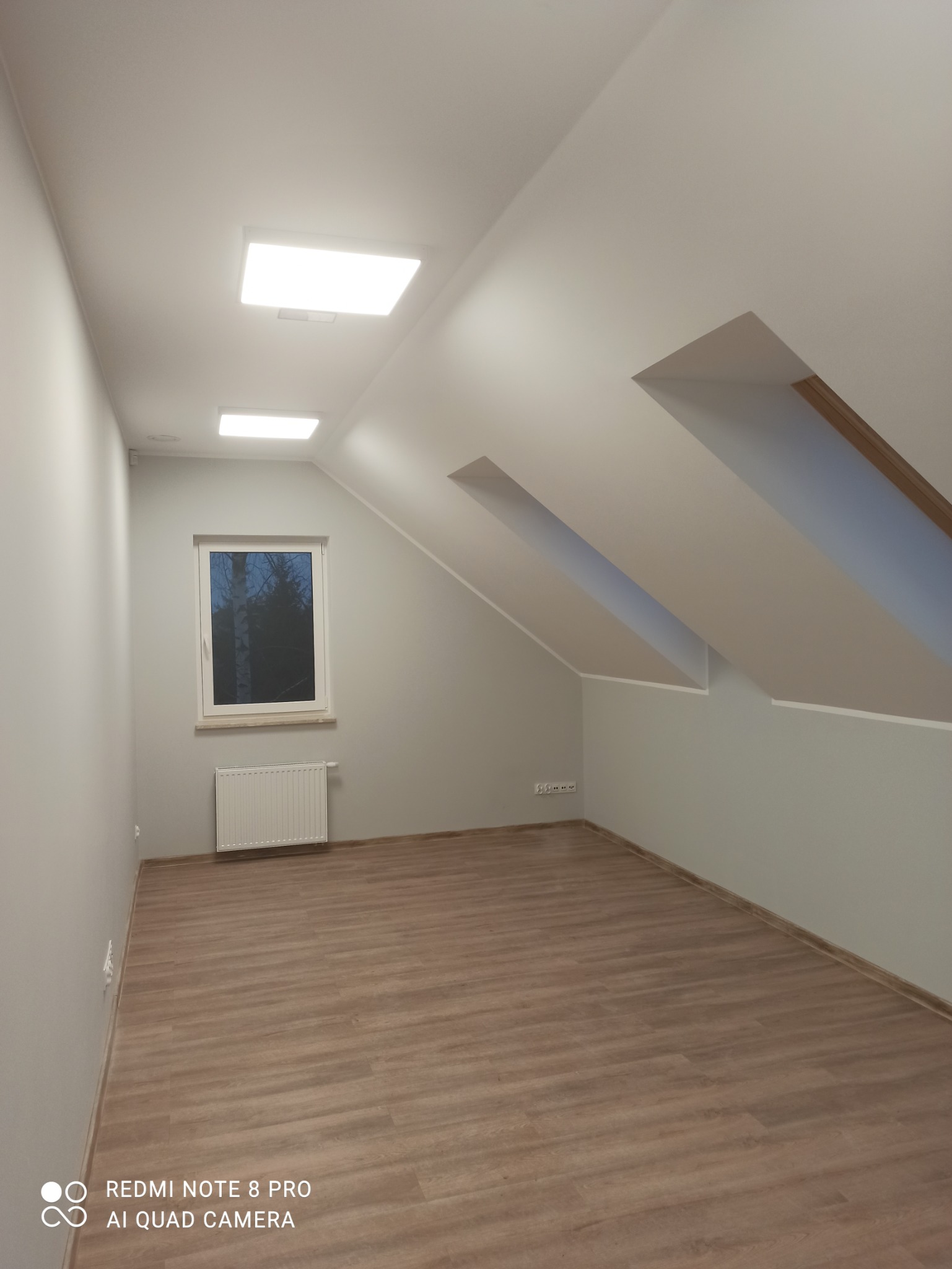 Poddasze po remoncie: świeżo pomalowane szare ściany, jasne panele podłogowe, biała rama okienna z widokiem na drzewa, grzejnik pod oknem, wbudowane kwadratowe lampy sufitowe.