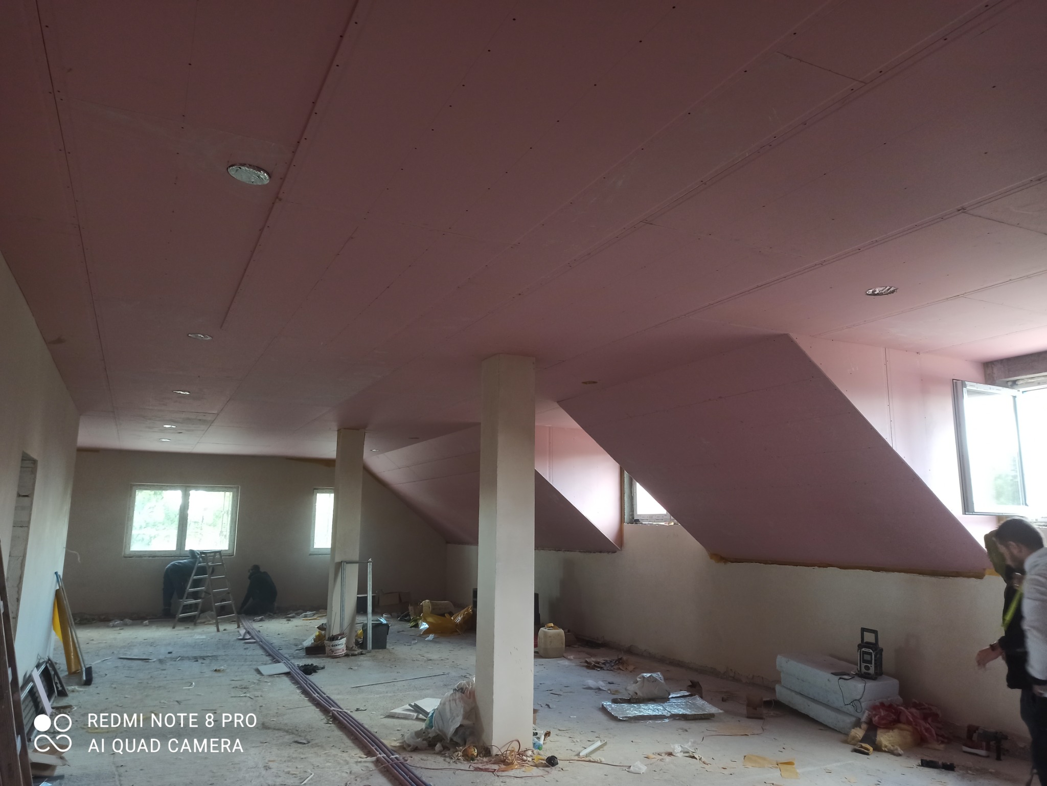 Montaż płyt na poddaszu, widoczne płyty gipsowo-kartonowe na skośnym suficie, pracownicy na drabinie i bałagan na podłodze.