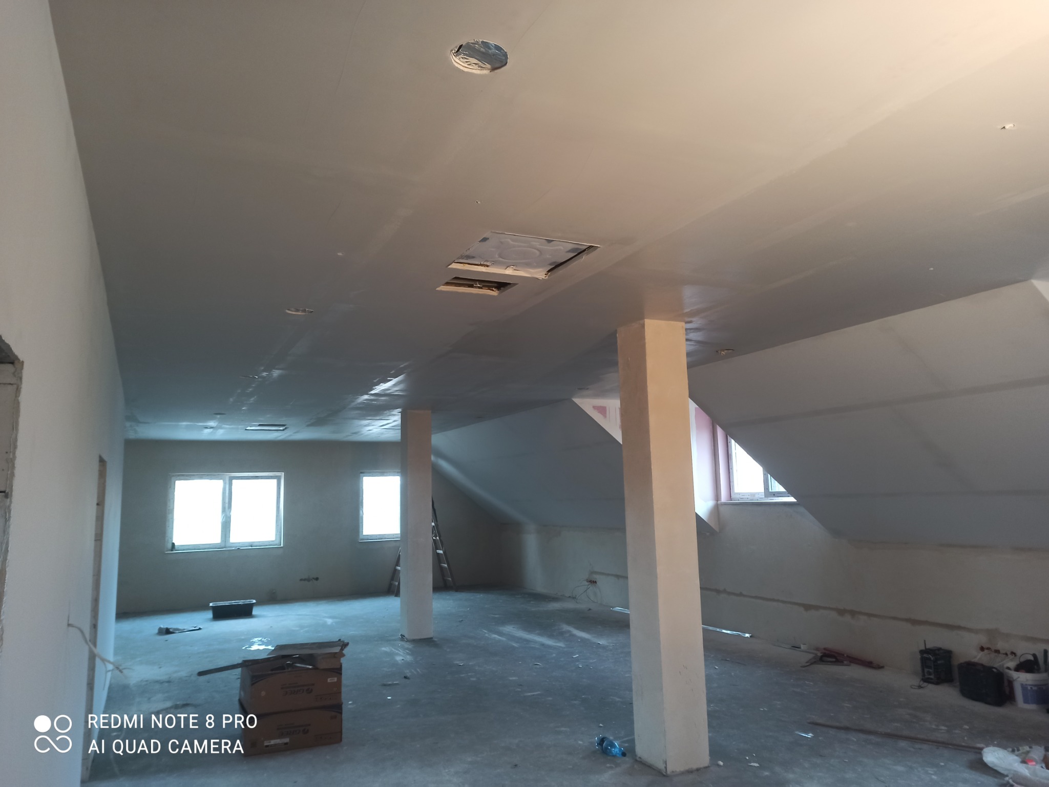 Poddasze w trakcie prac wykończeniowych: widoczne szpachlowane ściany i sufit z otworami na oświetlenie, podłoga zakryta folią ochronną, narzędzia i materiały budowlane w tle.