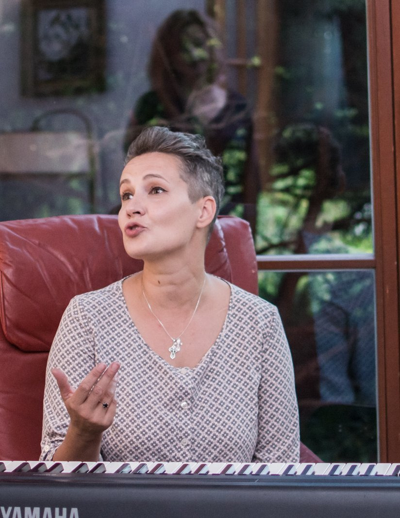 Kobieta z krótkimi włosami w bluzce w geometryczne wzory, siedząca przy keyboardzie Yamaha, gestykulująca podczas mówienia, w tle widoczne okno z zielenią i odbijającą się postacią.