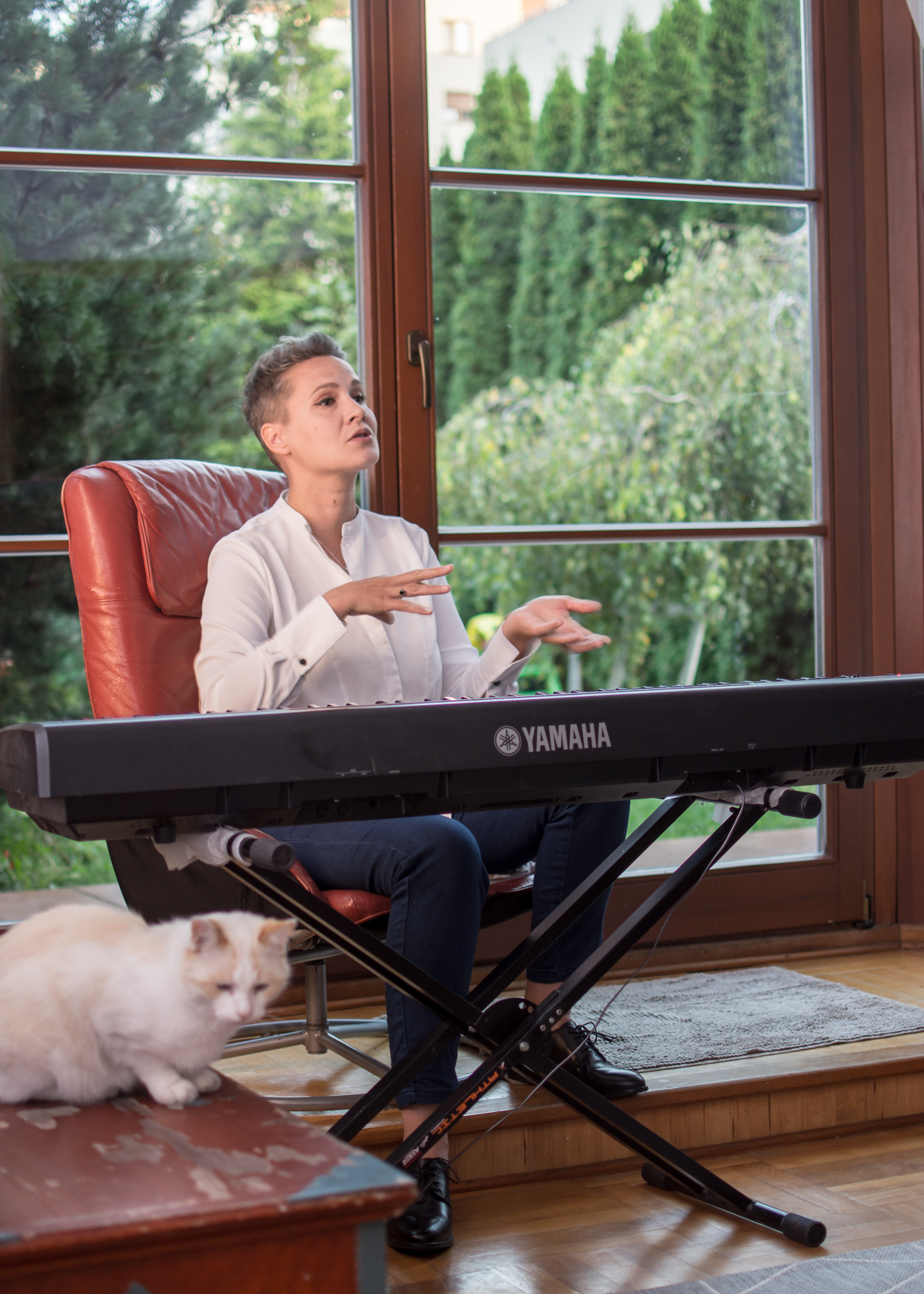 Kobieta w białej koszuli siedzi na czerwonym skórzanym krześle przed keyboardem Yamaha, gestykulując podczas lekcji, z białym kotem siedzącym na stoliku obok.