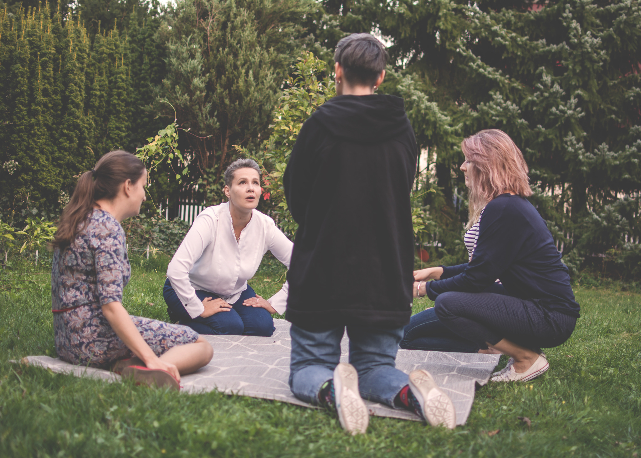 Grupa kobiet siedzi na kocu na trawie podczas zajęć na świeżym powietrzu, prawdopodobnie warsztatów interpersonalnych.