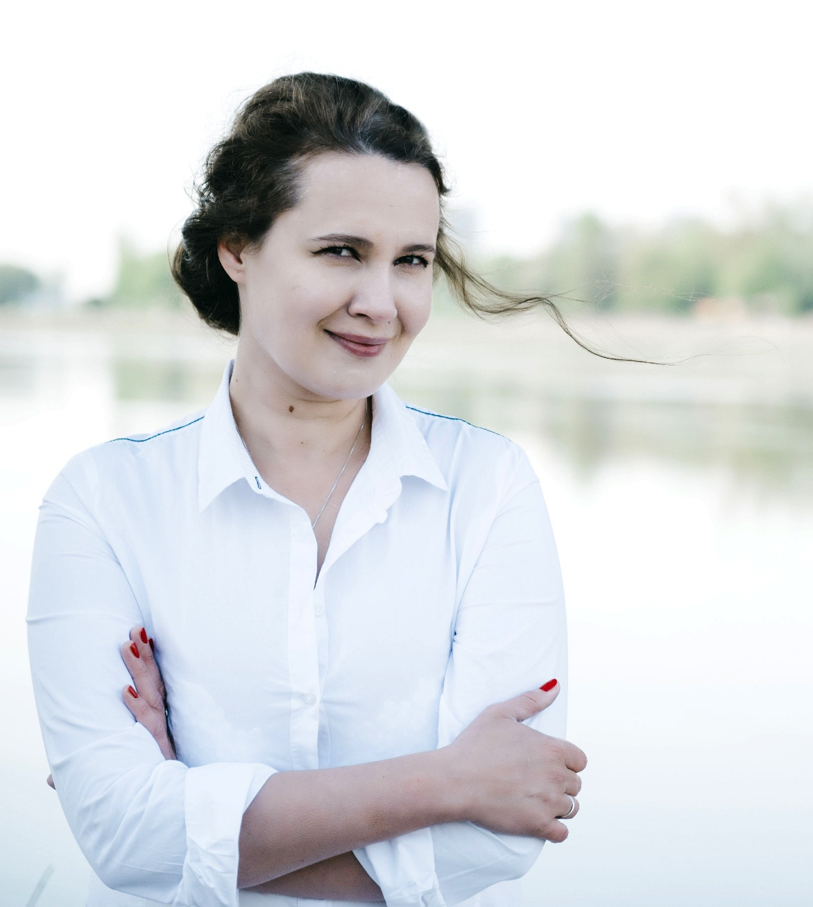 Portret kobiety w białej koszuli z założonymi rękami na tle rozmytego krajobrazu wodnego i drzew, z uśmiechem i kosmykiem włosów na wietrze.