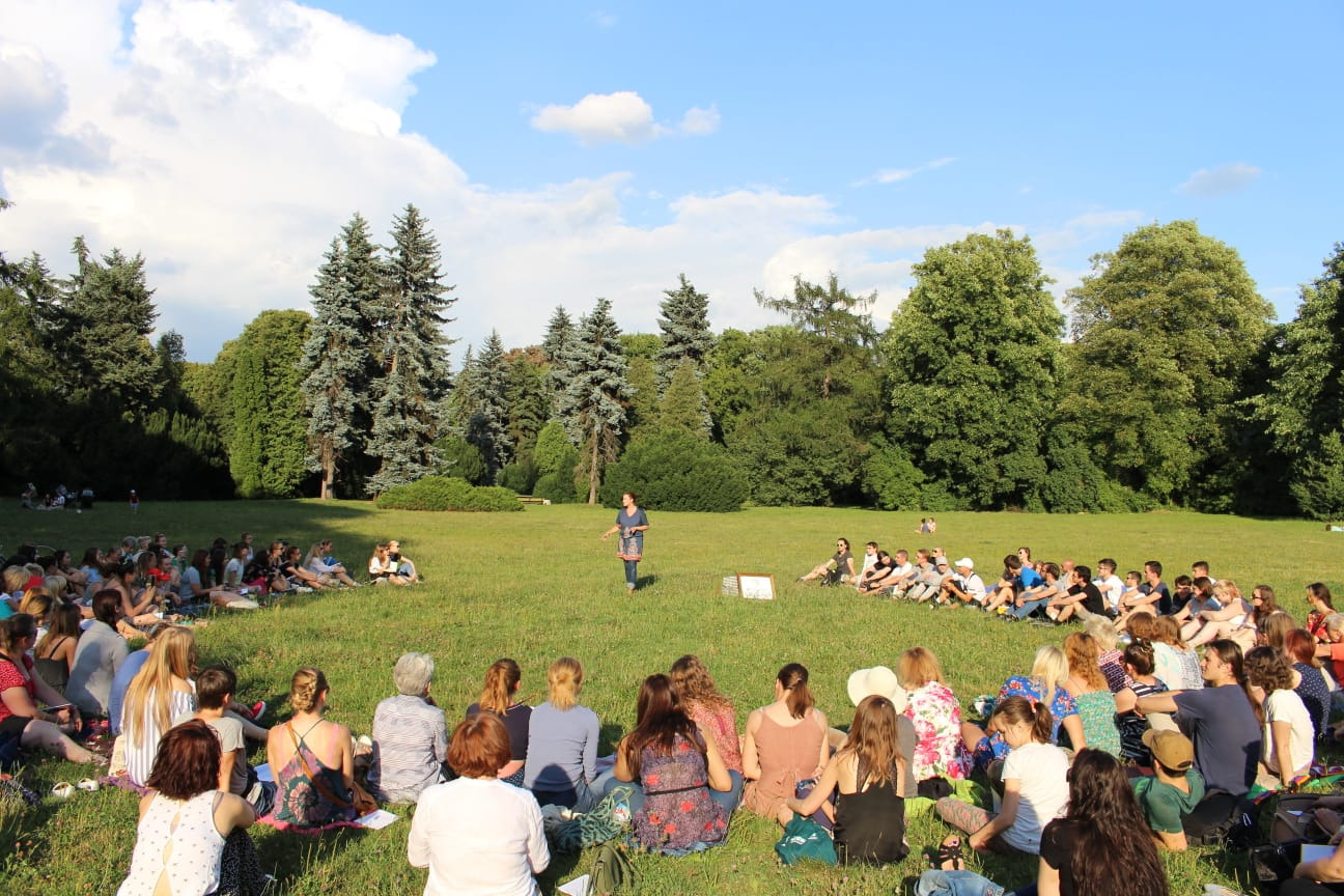 Grupa osób siedzi na trawie w parku, tworząc okrąg wokół kobiety stojącej pośrodku, która prawdopodobnie prowadzi warsztaty lub prezentację, w tle drzewa i błękitne niebo z chmurami.
