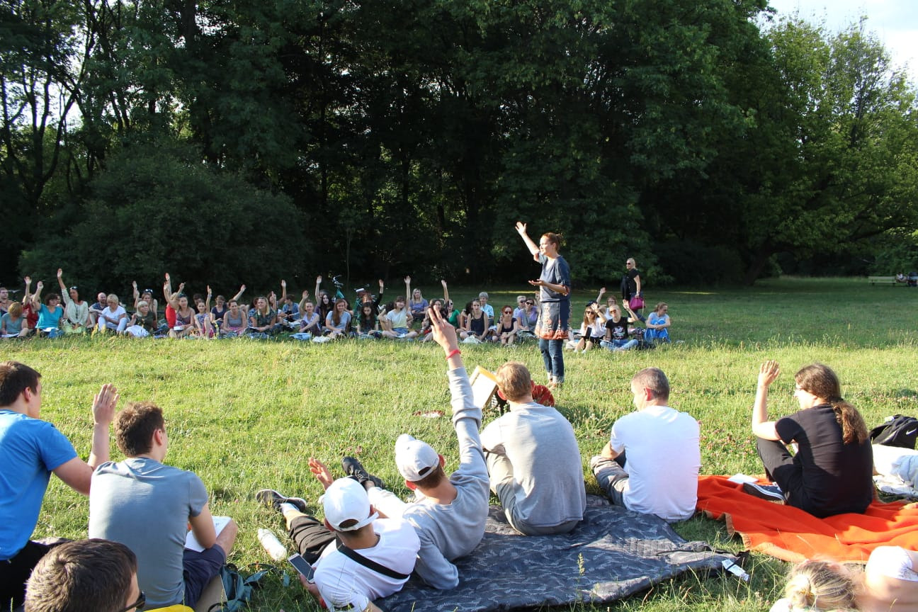 Szkolenie interpersonalne w plenerze: grupa osób siedzi na trawie, słuchając prelegentki z podniesioną ręką, w tle drzewa.