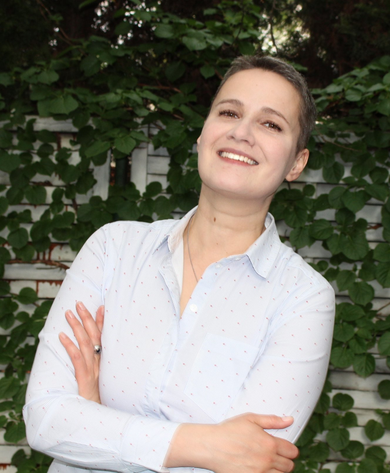 Uśmiechnięta kobieta o krótkich włosach, ubrana w błękitną koszulę w drobne czerwone kropeczki, stoi na tle białego płotu porośniętego zielonym bluszczem. Ręce skrzyżowane, na palcu pierścionek.