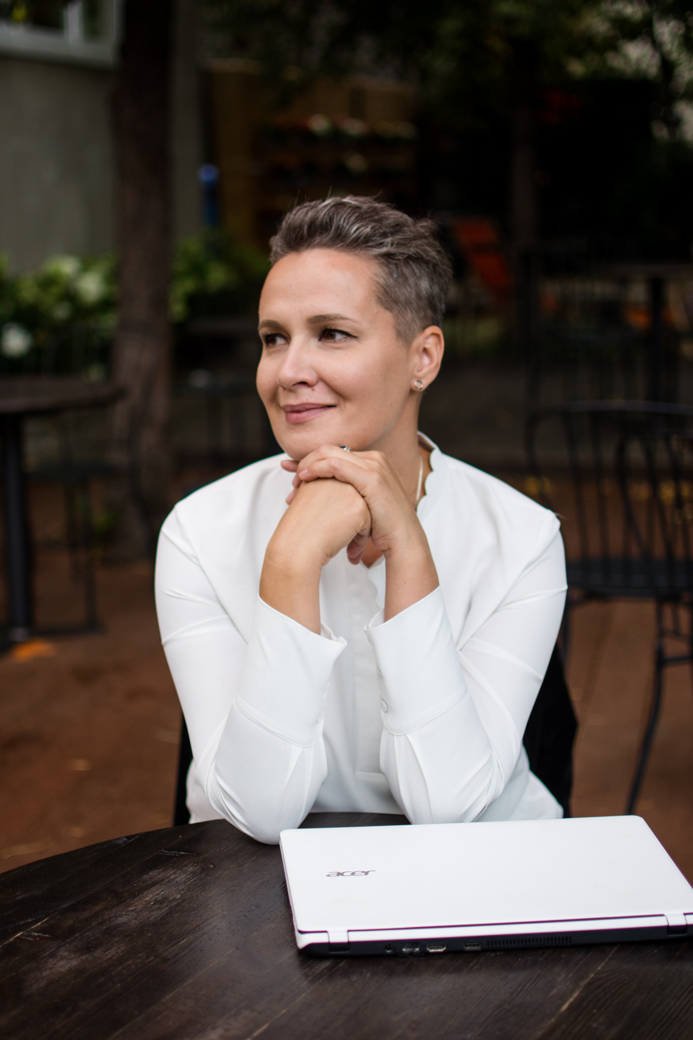 Kobieta w białej koszuli siedzi przy drewnianym stole z laptopem Acer, zamyślona i uśmiechnięta, w tle stoliki kawiarniane.