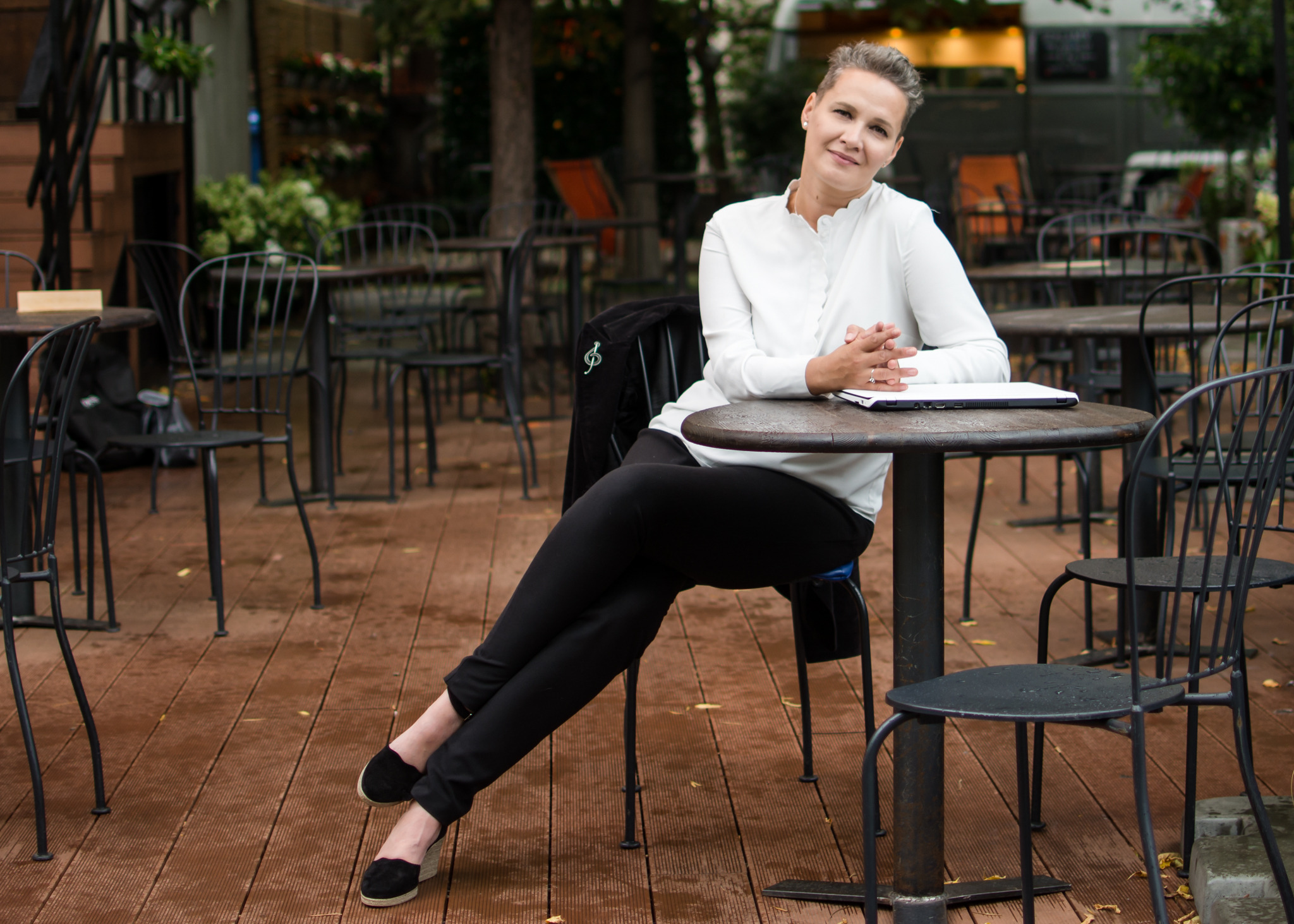 Kobieta w białej koszuli siedzi przy stoliku na zewnątrz, z laptopem, na drewnianym tarasie kawiarni, w tle widoczne stoliki i zieleń.