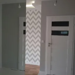 Nowoczesne wnętrze mieszkania z szaro-białą tapetą w zygzaki, białe drzwi z czarnymi akcentami i podłogą z paneli, widok z korytarza.