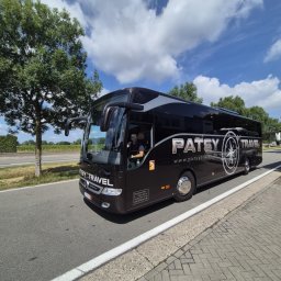 M-TRANS - Czarny autokar turystyczny Patsy Travel z kierowcą w okularach przeciwsłonecznych, zaparkowany na poboczu drogi w słoneczny dzień. Widoczne logo firmy i dane kontaktowe.