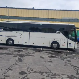 Biały autokar marki Setra z logo Polbus zaparkowany na placu z popękanym asfaltem, na tle żółtego budynku. Widoczny fragment innego pojazdu i kontener.