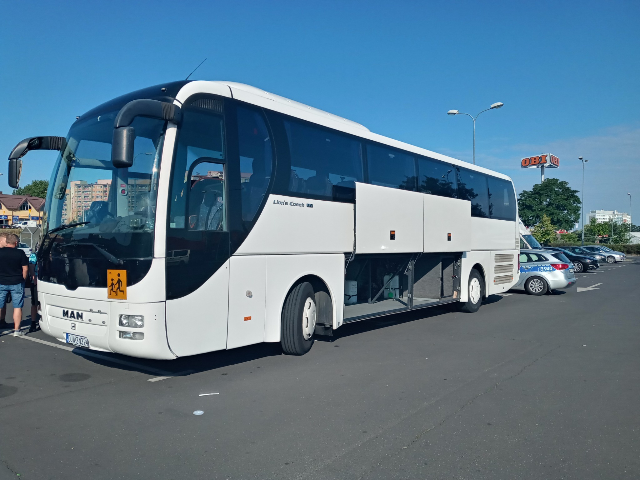 Biały autokar MAN Lion's Coach z otwartymi lukami bagażowymi na parkingu, z widocznym znakiem 'dzieci' na przedniej szybie i zaparkowanymi samochodami w tle.