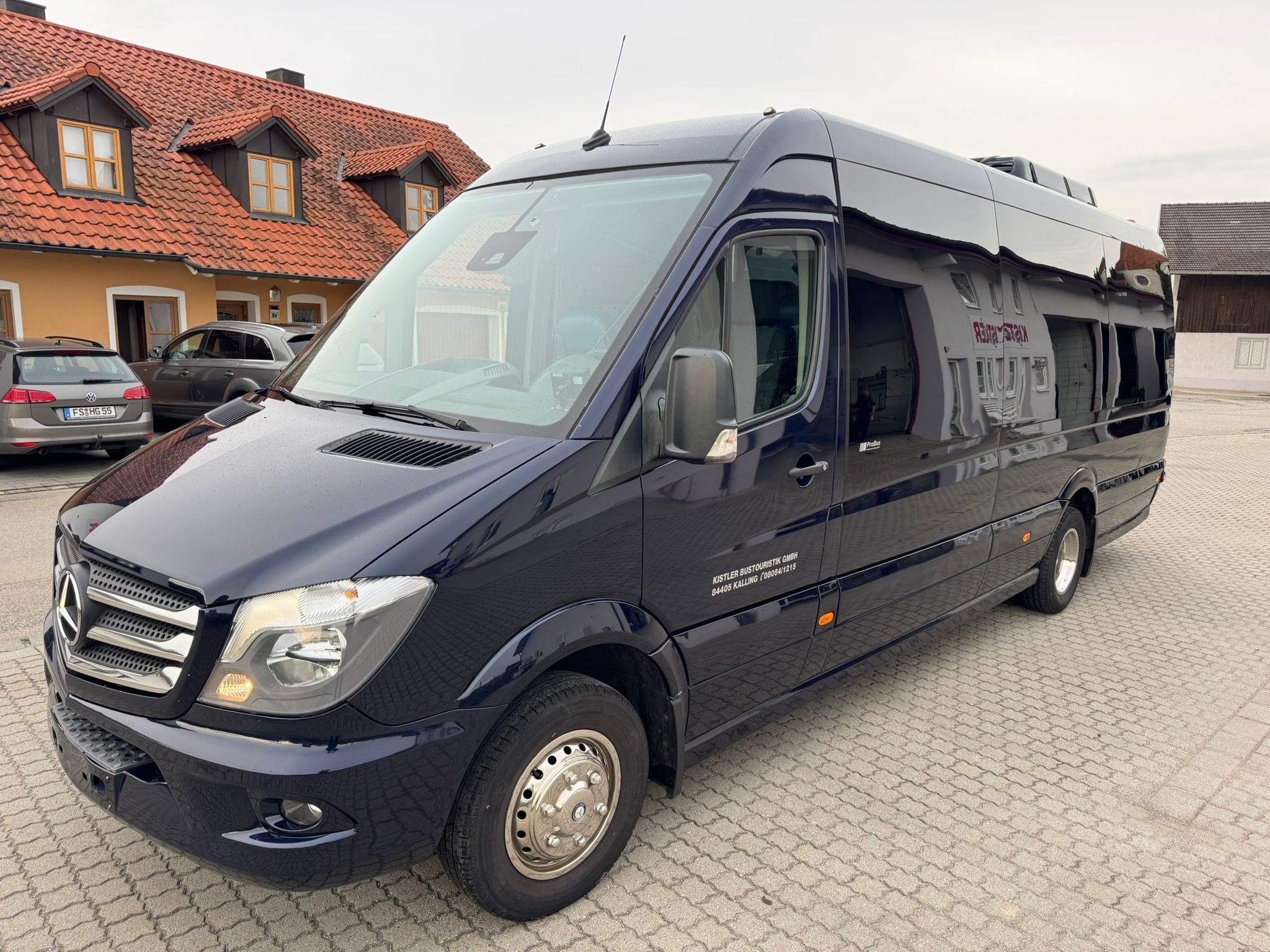 Elegancki, granatowy bus Mercedes-Benz Sprinter z chromowanymi felgami na brukowanym placu, z budynkiem w tle. Widoczny napis 'KISTLER BUS TOURISTIK'.