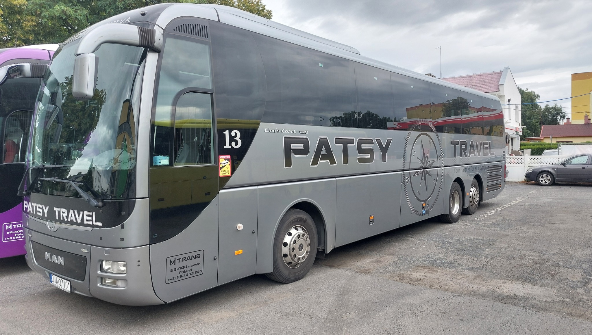 Szary autokar turystyczny Patsy Travel z numerem 13, zaparkowany na zewnątrz, z widocznymi detalami marki MAN i oznaczeniami MTRANS. W tle budynki i inne pojazdy.