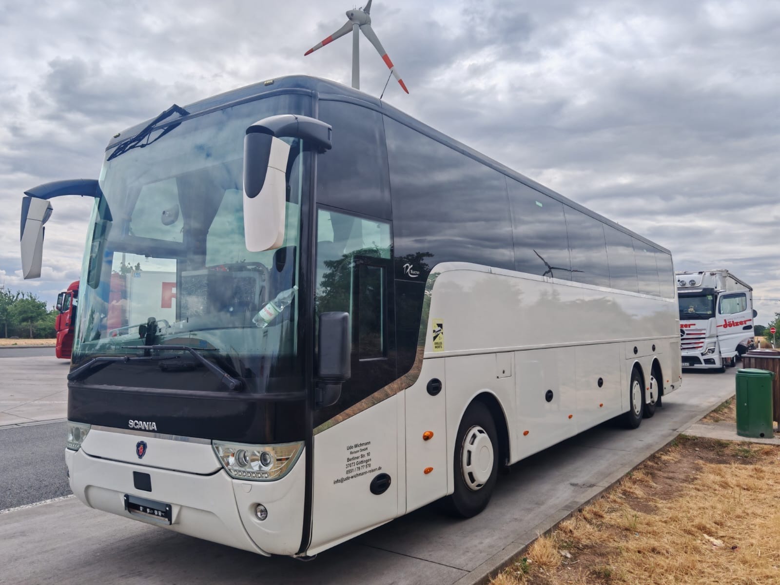 Elegancki, biało-czarny autokar Scania z turbiną w tle, zaparkowany na betonowym placu. Widoczne logo firmy Udo Wichmann i częściowo zaparkowana ciężarówka.