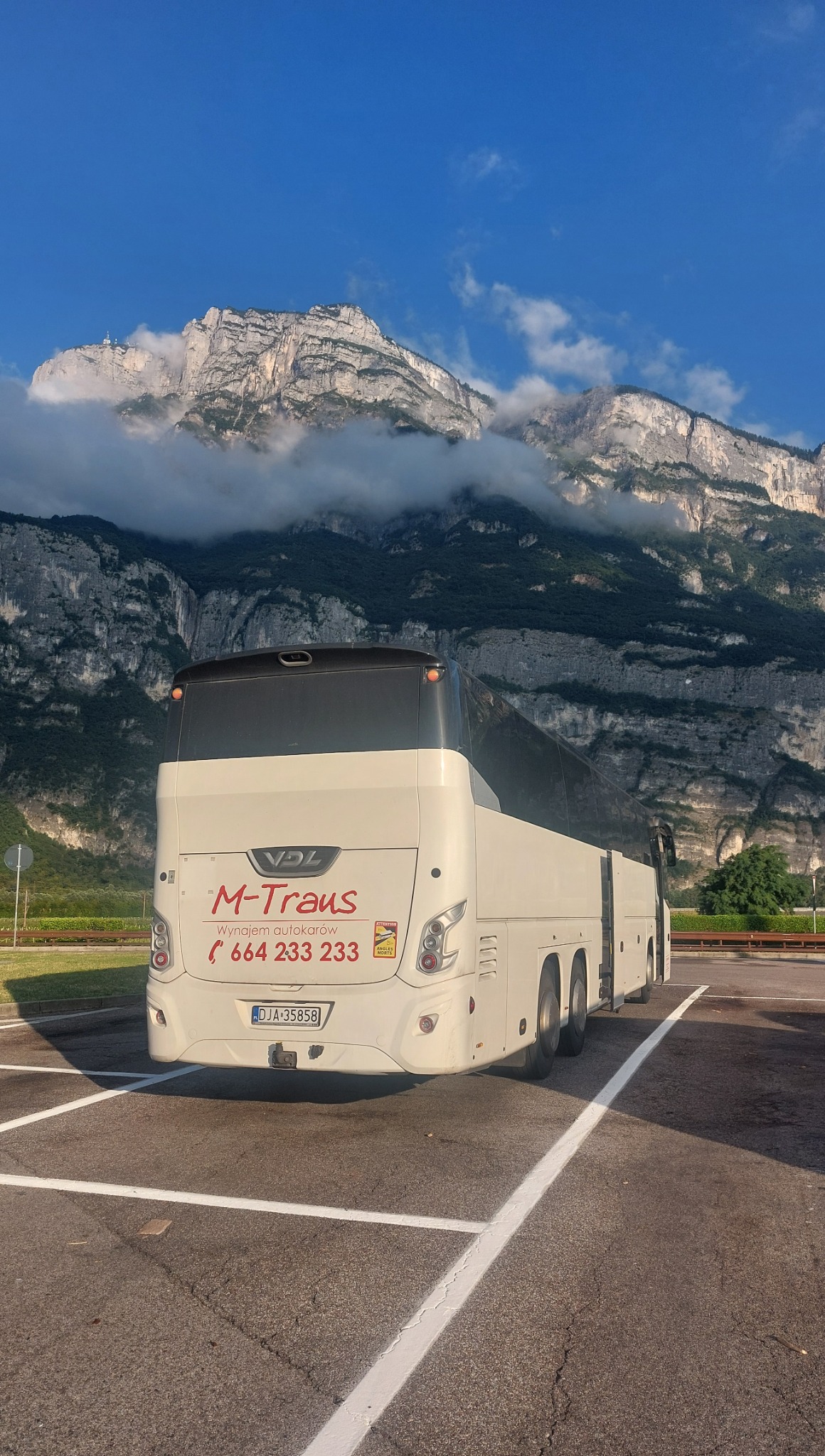 Autokar M-Trans na tle gór z chmurami, zaparkowany na parkingu. Widoczny numer telefonu i napis 'Wynajem autokarów'.