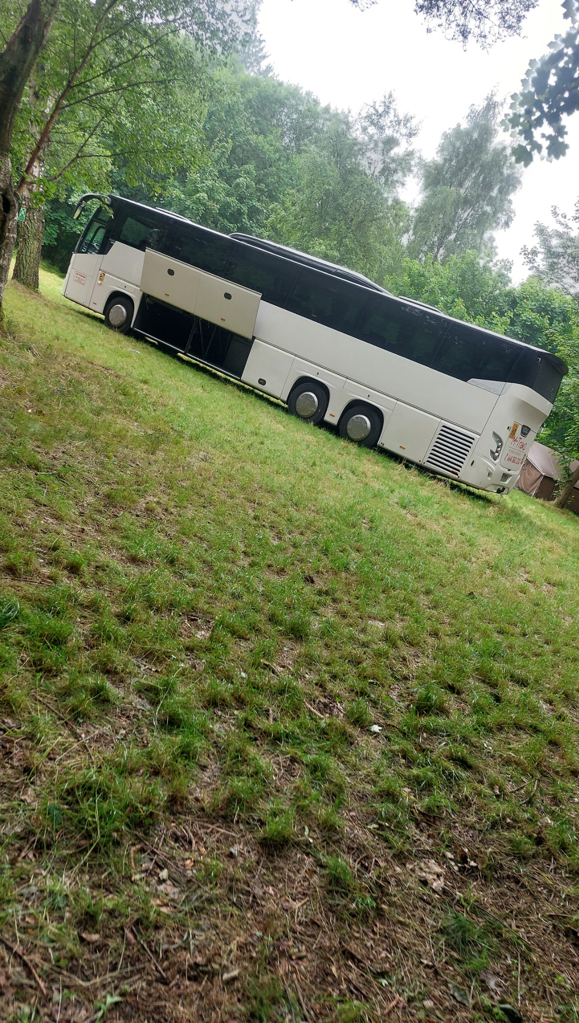 Biały autokar na trawiastym zboczu, widoczny z boku, z otwartymi drzwiami w środkowej części. W tle drzewa i krzewy, co sugeruje postój na łonie natury.