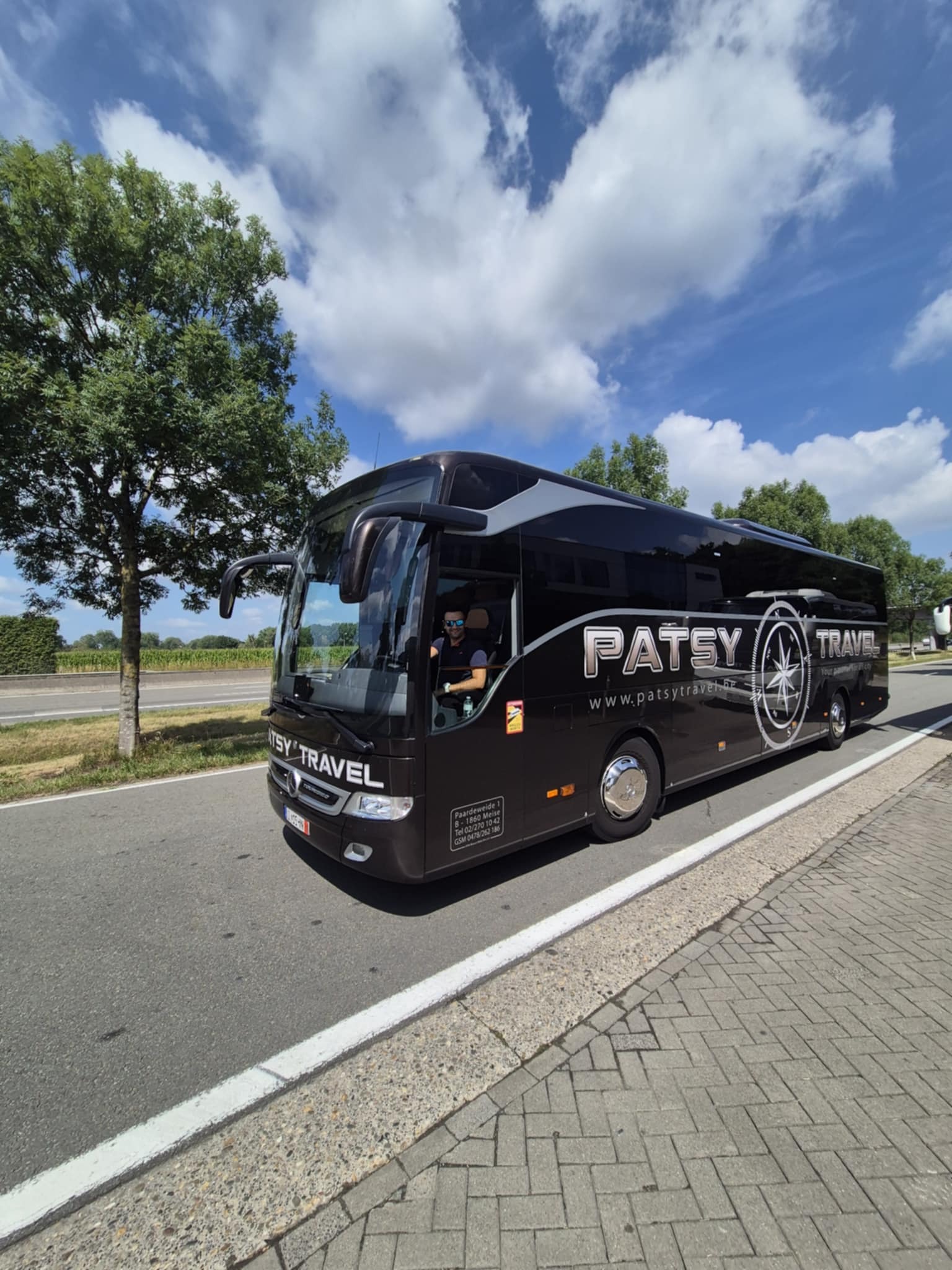 Czarny autokar turystyczny Patsy Travel z kierowcą w okularach przeciwsłonecznych, zaparkowany na poboczu drogi w słoneczny dzień. Widoczne logo firmy i dane kontaktowe.