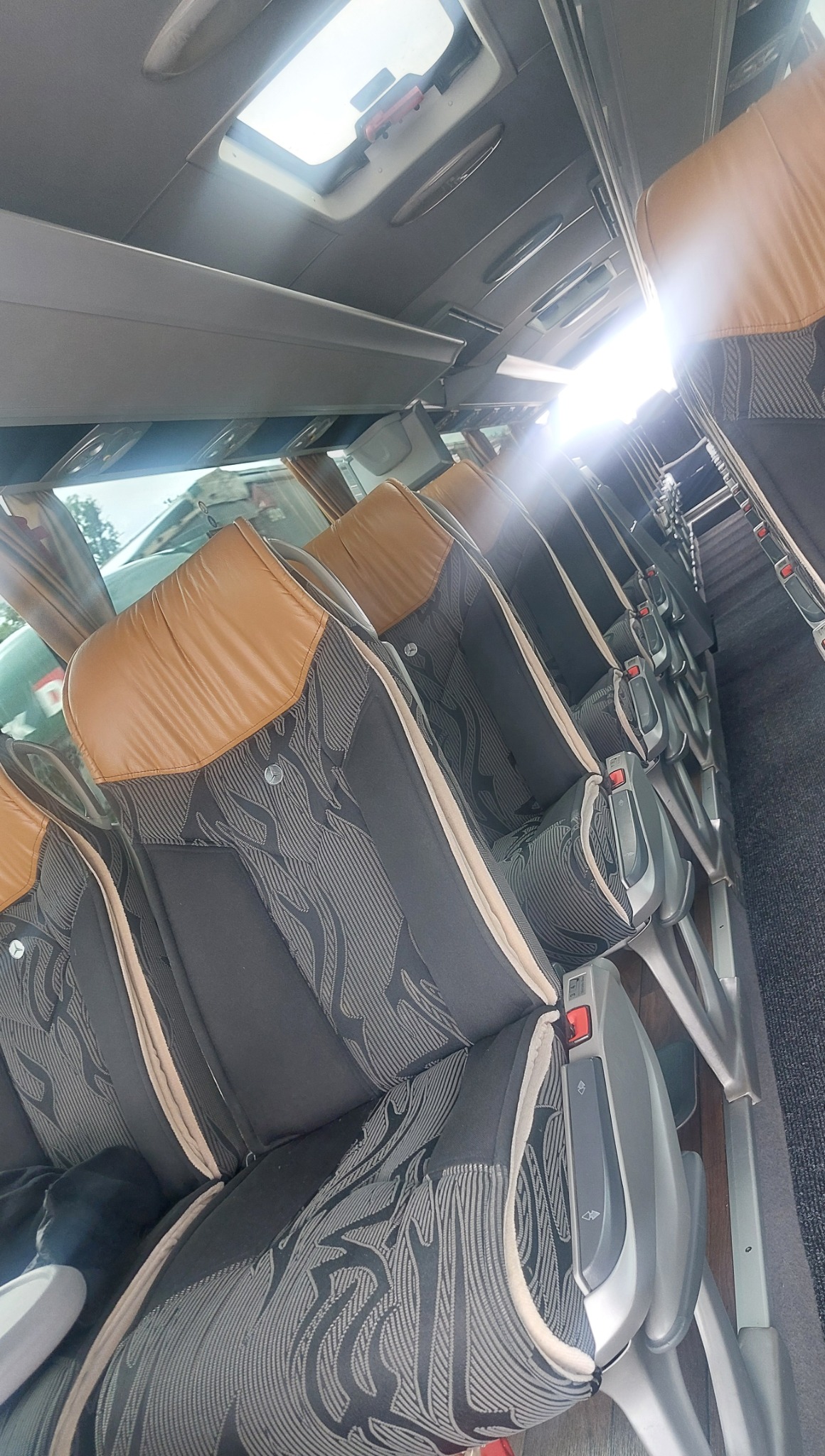 Wnętrze autobusu turystycznego: rzędy foteli z brązowymi zagłówkami i wzorzystą tapicerką, widok z boku, oświetlenie słoneczne wpadające przez okna dachowe.