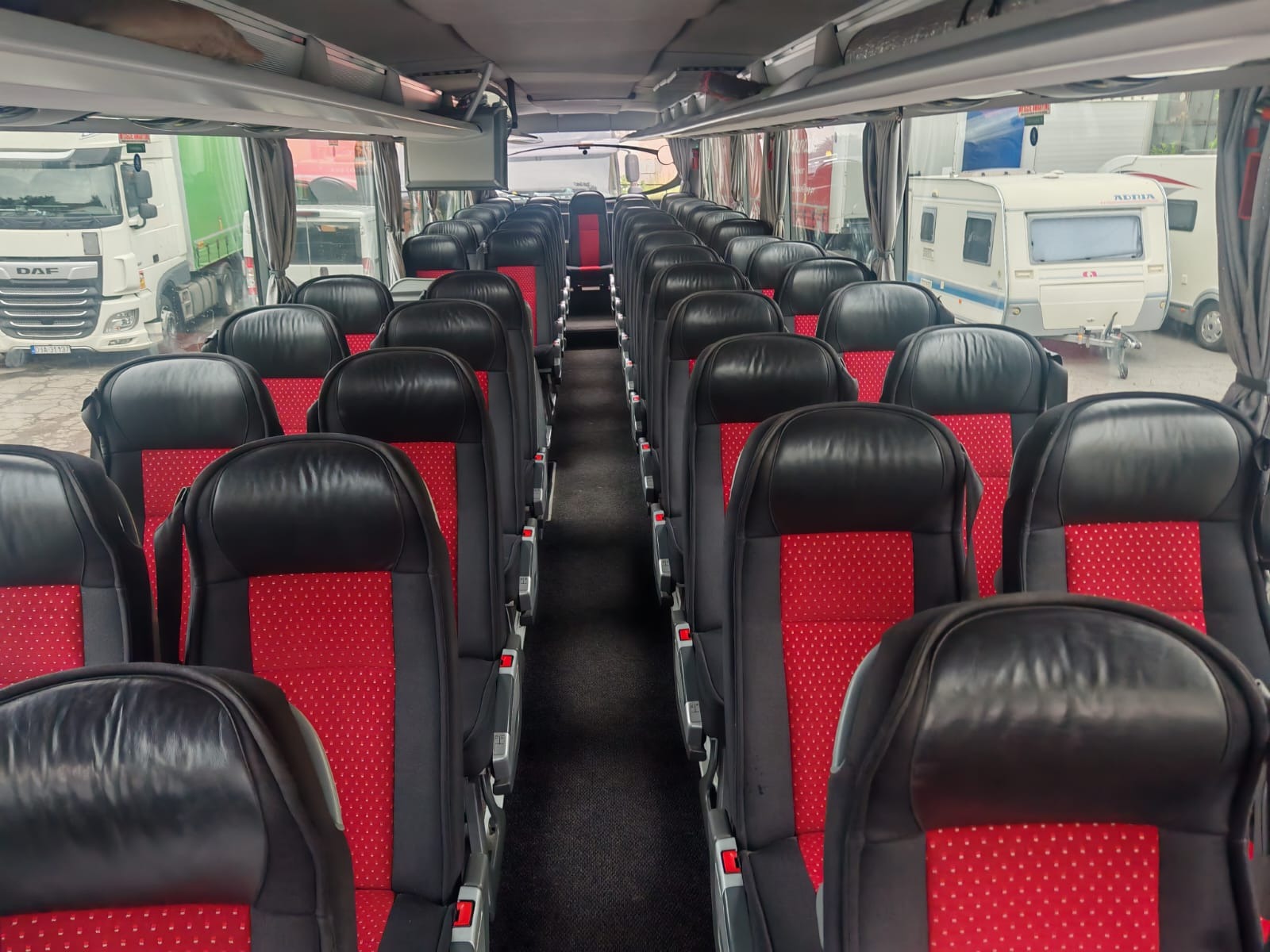 Wnętrze autobusu turystycznego z rzędami czarno-czerwonych foteli. Za oknami widoczne ciężarówki i przyczepy kempingowe.