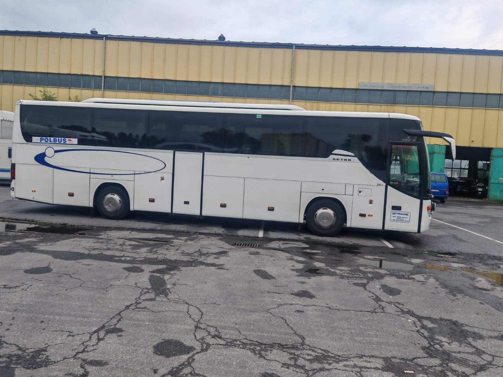 Biały autokar marki Setra z logo Polbus zaparkowany na placu z popękanym asfaltem, na tle żółtego budynku. Widoczny fragment innego pojazdu i kontener.