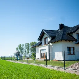 Nowoczesny dom jednorodzinny z czarnym dachem i ogrodzeniem panelowym na tle zielonego pola i błękitnego nieba.