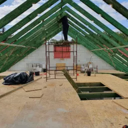 Konstrukcja dachu z zielonymi belkami, widok od wewnątrz, pracownik na rusztowaniu mocuje elementy, w tle ściana z białych bloczków i fragment dachu sąsiedniego budynku.