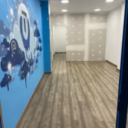 Wnętrze biura w trakcie prac wykończeniowych: świeżo położona podłoga z paneli imitujących drewno, pomalowana na niebiesko ściana z dekoracyjnym motywem chmur i logo, ściany przygotowane...