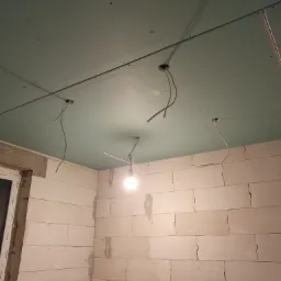 Surowy stan wnętrza: ściany z bloczków, sufit z płyt kartonowo-gipsowych z wystającymi przewodami elektrycznymi, tymczasowe oświetlenie żarówką.