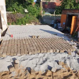 Układanie kostki brukowej na podsypce piaskowej, widoczny fragment ułożonej nawierzchni i przygotowane kostki, w tle budynek z cegły i zieleń.