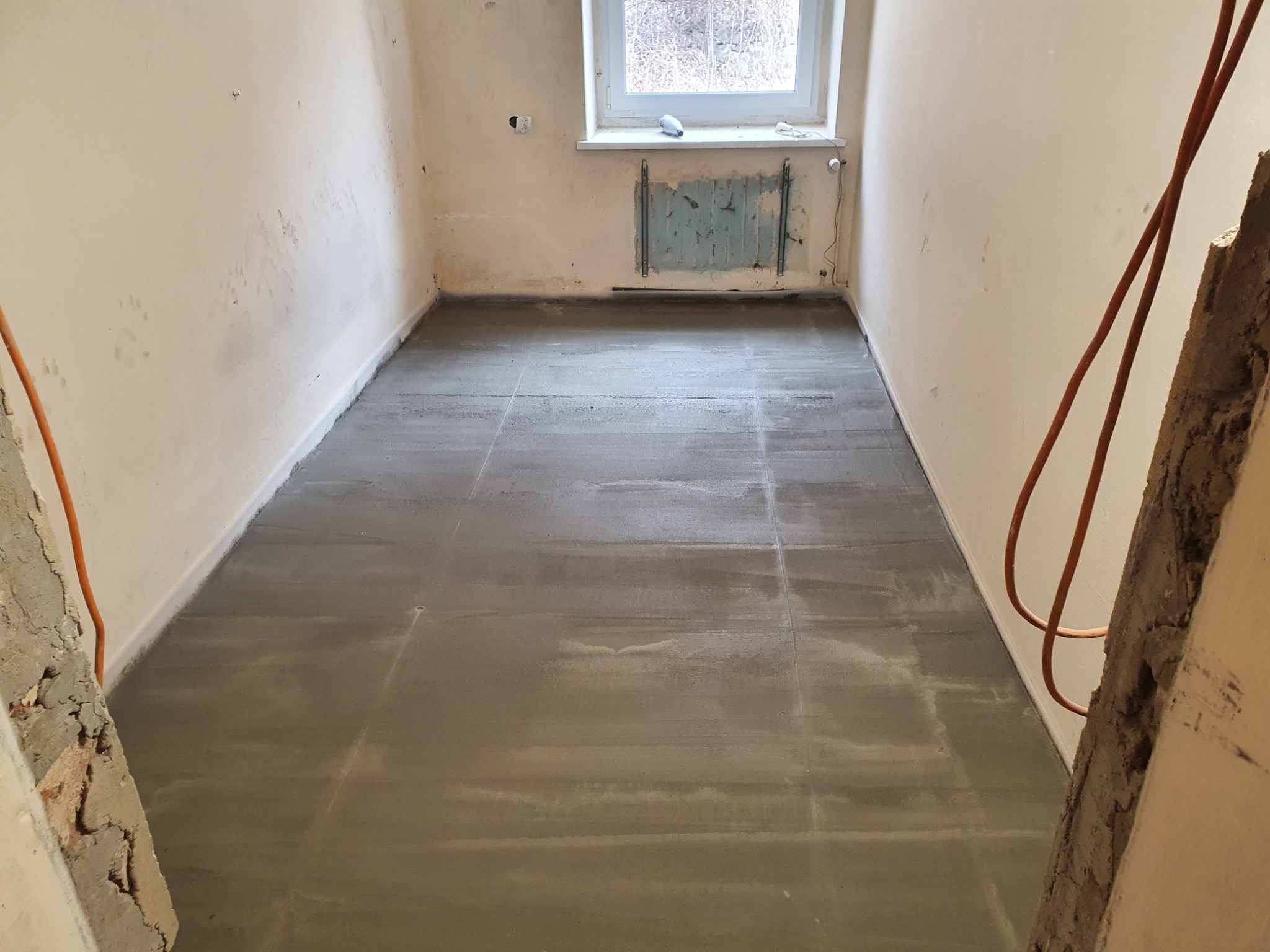 Wylany, świeży podkład podłogowy w pomieszczeniu z oknem i starym grzejnikiem, widoczne fragmenty ścian przed wykończeniem.