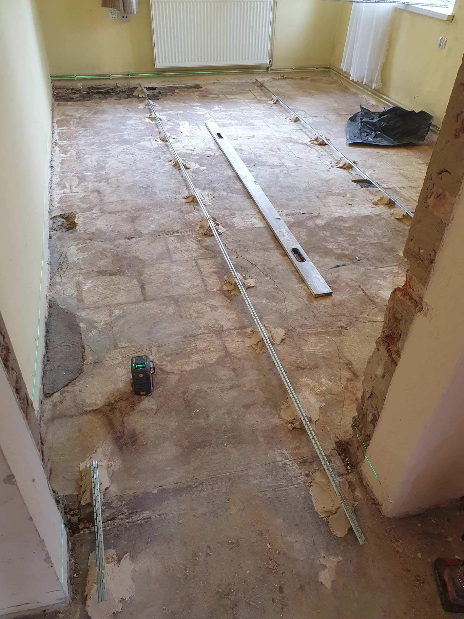 Przygotowanie podłoża pod nową podłogę w pokoju: widoczne listwy poziomujące, poziomica, laser krzyżowy i resztki starej posadzki.