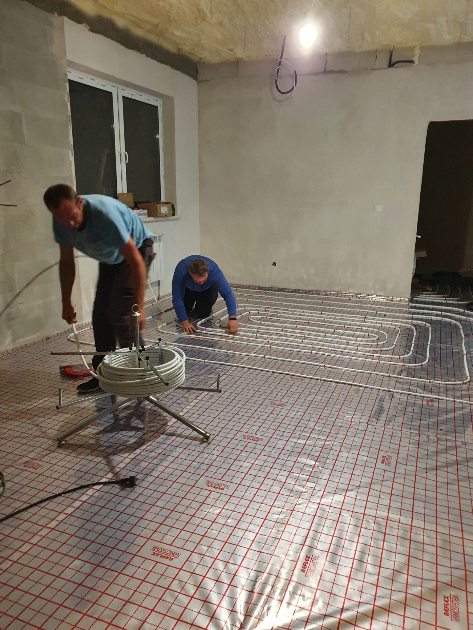 Instalacja ogrzewania podłogowego: dwóch mężczyzn układa białe rury na folii z czerwoną siatką, w surowym wnętrzu z widoczną izolacją na suficie i oknem w tle.