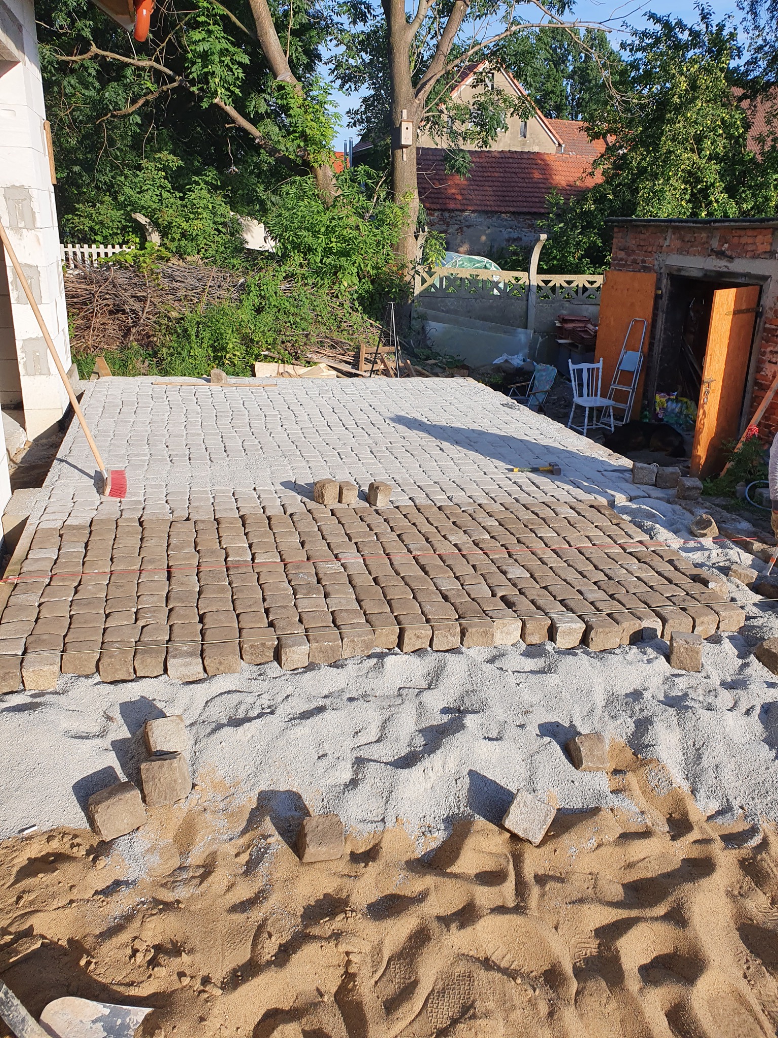 Układanie kostki brukowej na podsypce piaskowej, widoczny fragment ułożonej nawierzchni i przygotowane kostki, w tle budynek z cegły i zieleń.