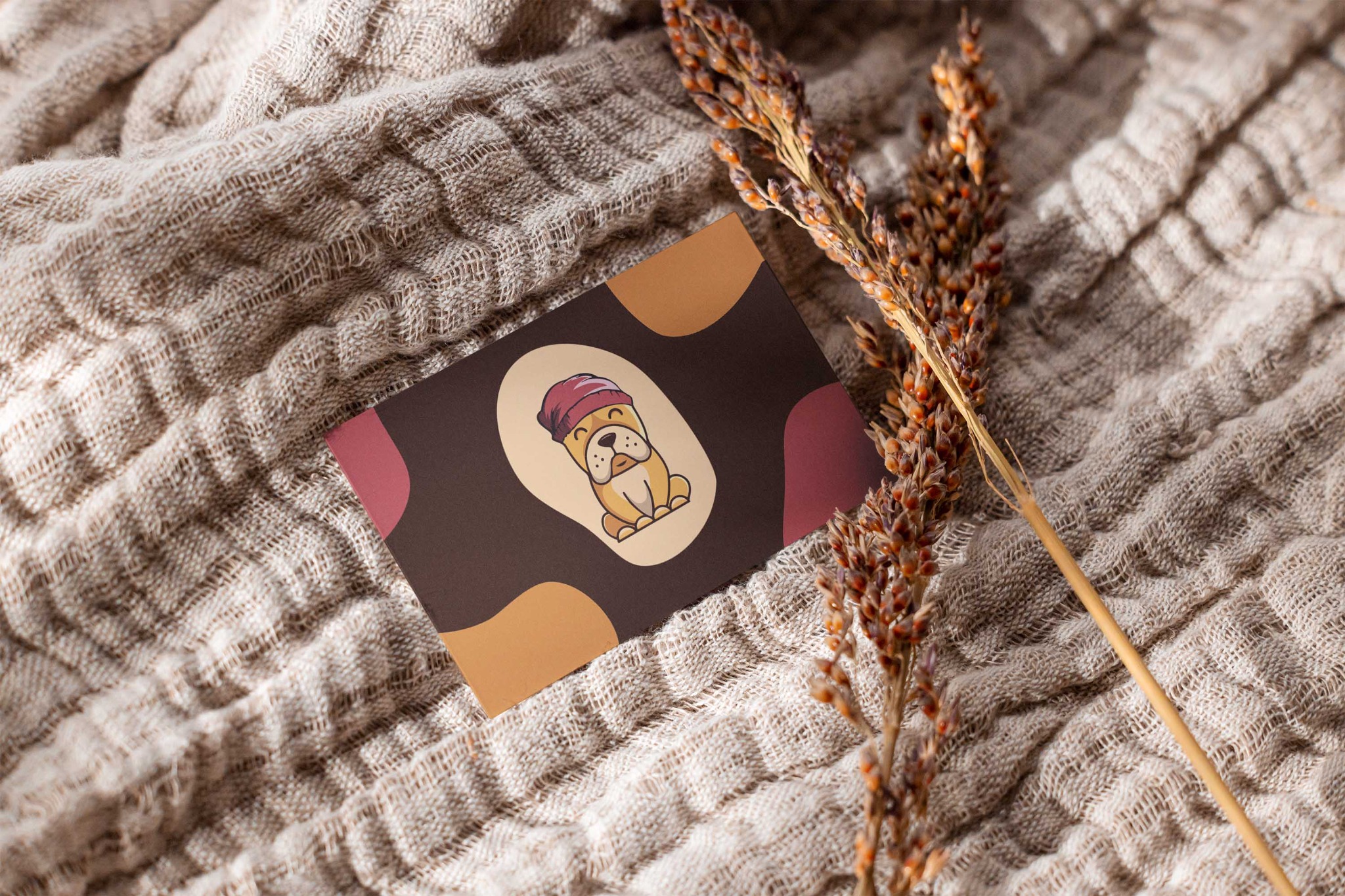 Karta z ilustracją buldoga francuskiego w czapce, leżąca na beżowej, teksturowanej tkaninie obok gałązki z brązowymi kłosami.