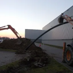 Dwa pomarańczowe traktory z łyżkami układają czarną, grubą rurę w wykopie ziemnym na tle budynku przemysłowego o blaszanej elewacji, widoczne znaki ostrzegawcze i zaparkowane samochody w oddali...