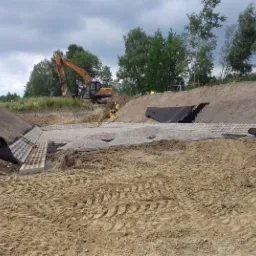 Budowa przepustu drogowego: Wykop z ułożonymi prefabrykatami betonowymi, warstwą żwiru i czarną geowłókniną, w tle koparka i pracownicy w żółtych kamizelkach.